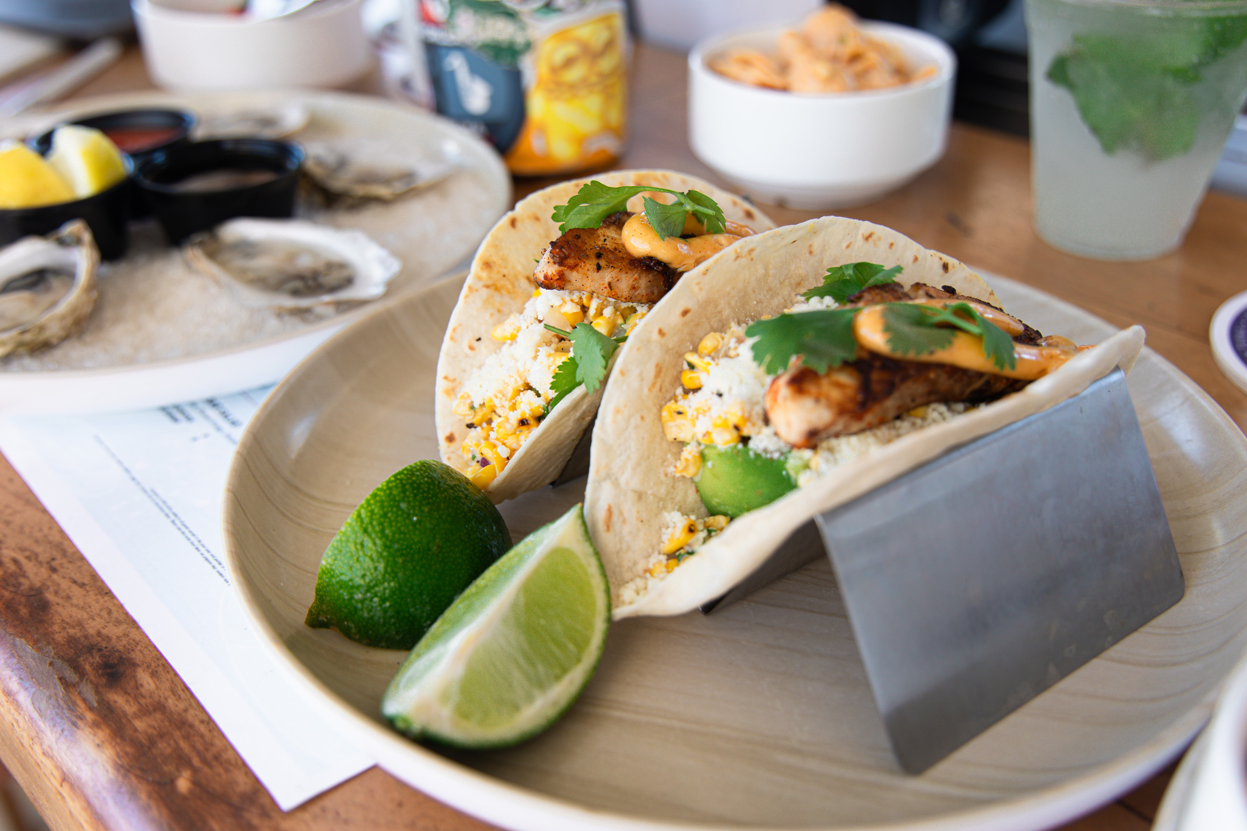 Two fish tacos with lime wedges and oysters in background.