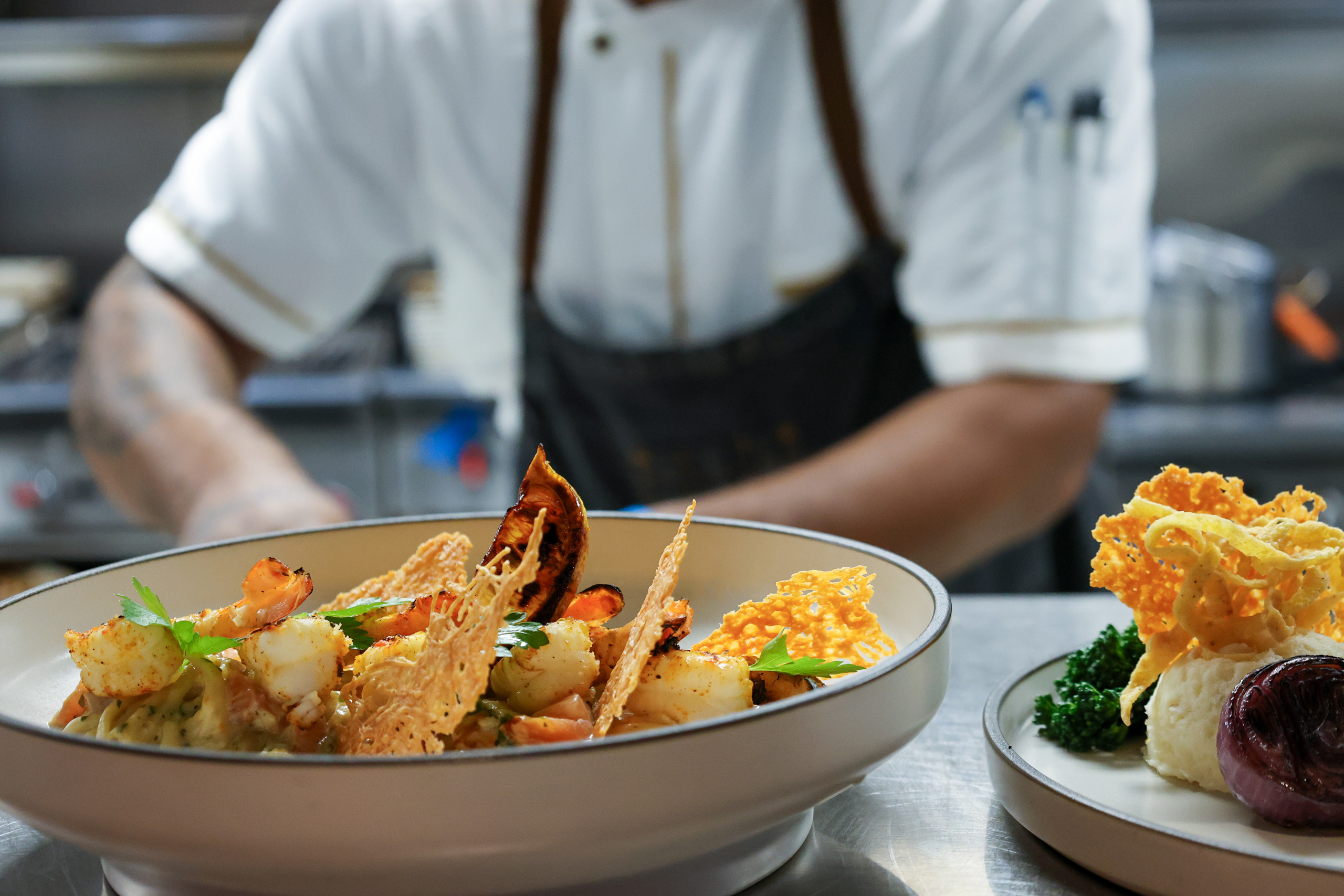 Chef plating seafood pasta with crispy garnishes in kitchen.