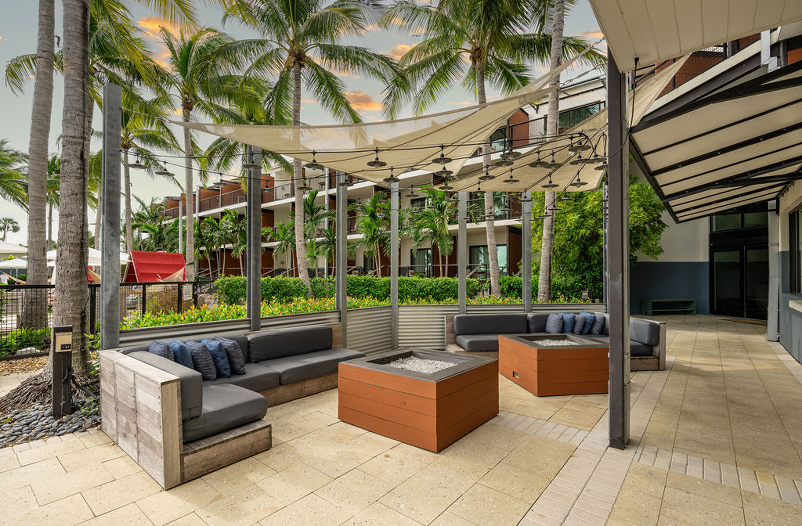 Outdoor lounge with shaded seating and fire pits among palm trees.