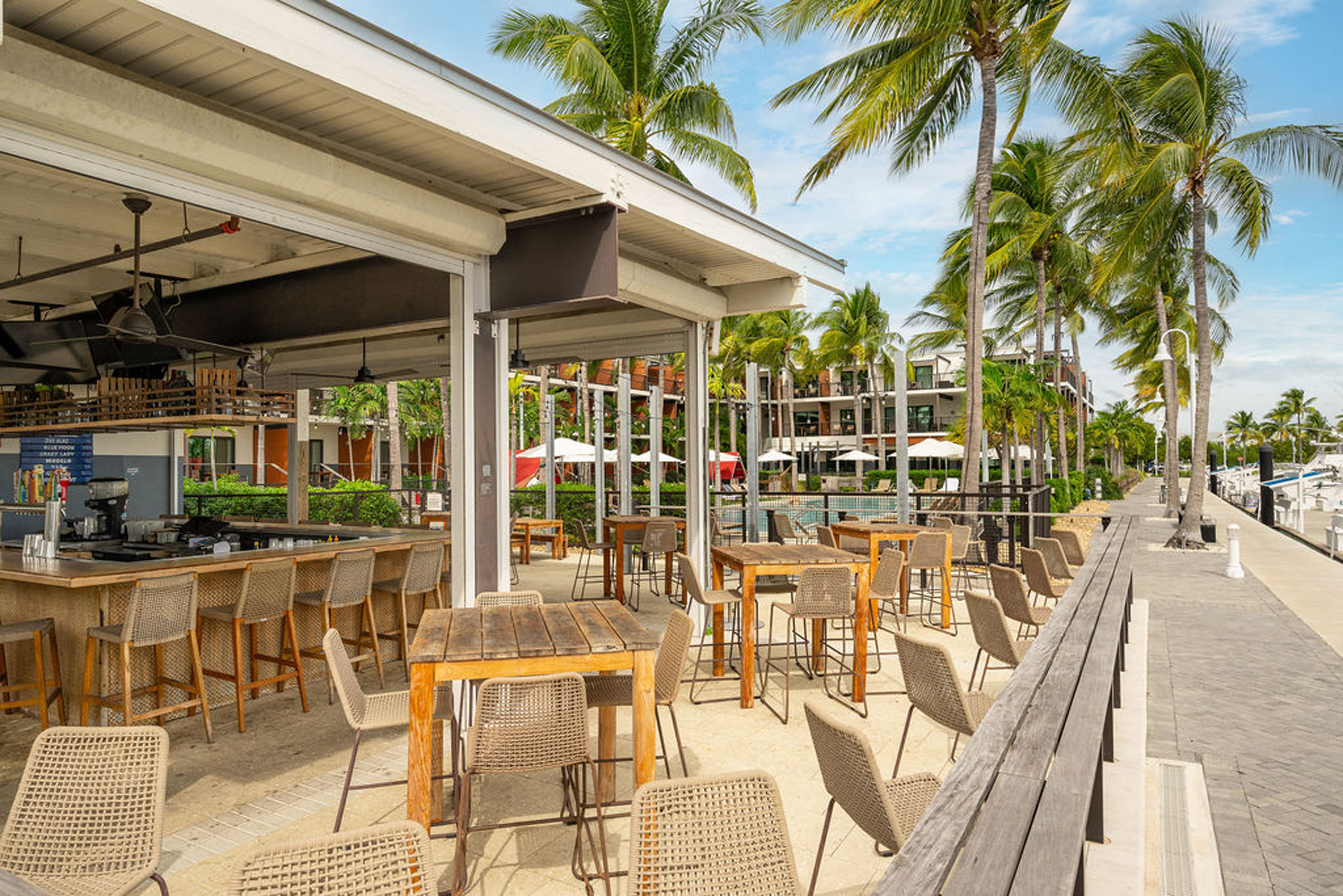 Waterfront bar with high-top tables and stools under palm trees.