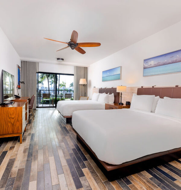 Spacious room with two queen beds, wood floors, desk, TV, and balcony access overlooking the pool.