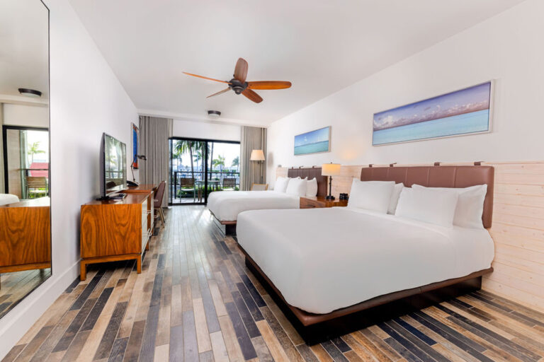 Spacious room with two queen beds, wood floors, desk, TV, and balcony access overlooking the pool.