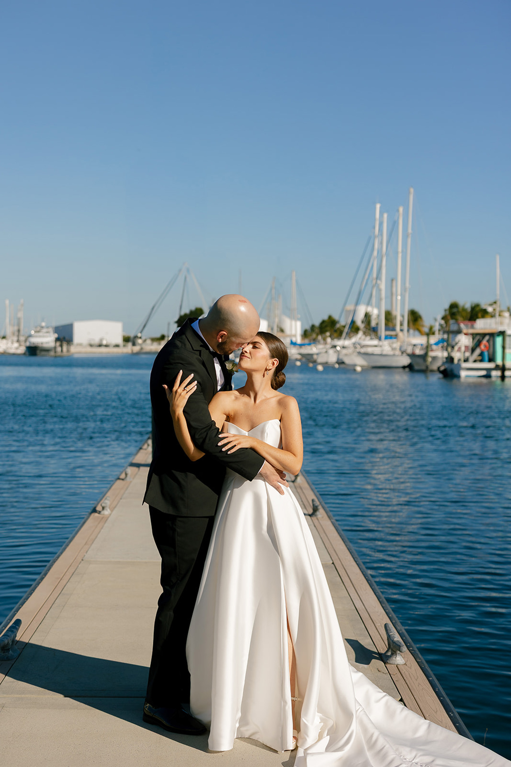 Bride and groom kiss on marina dock surrounded by sailboats.