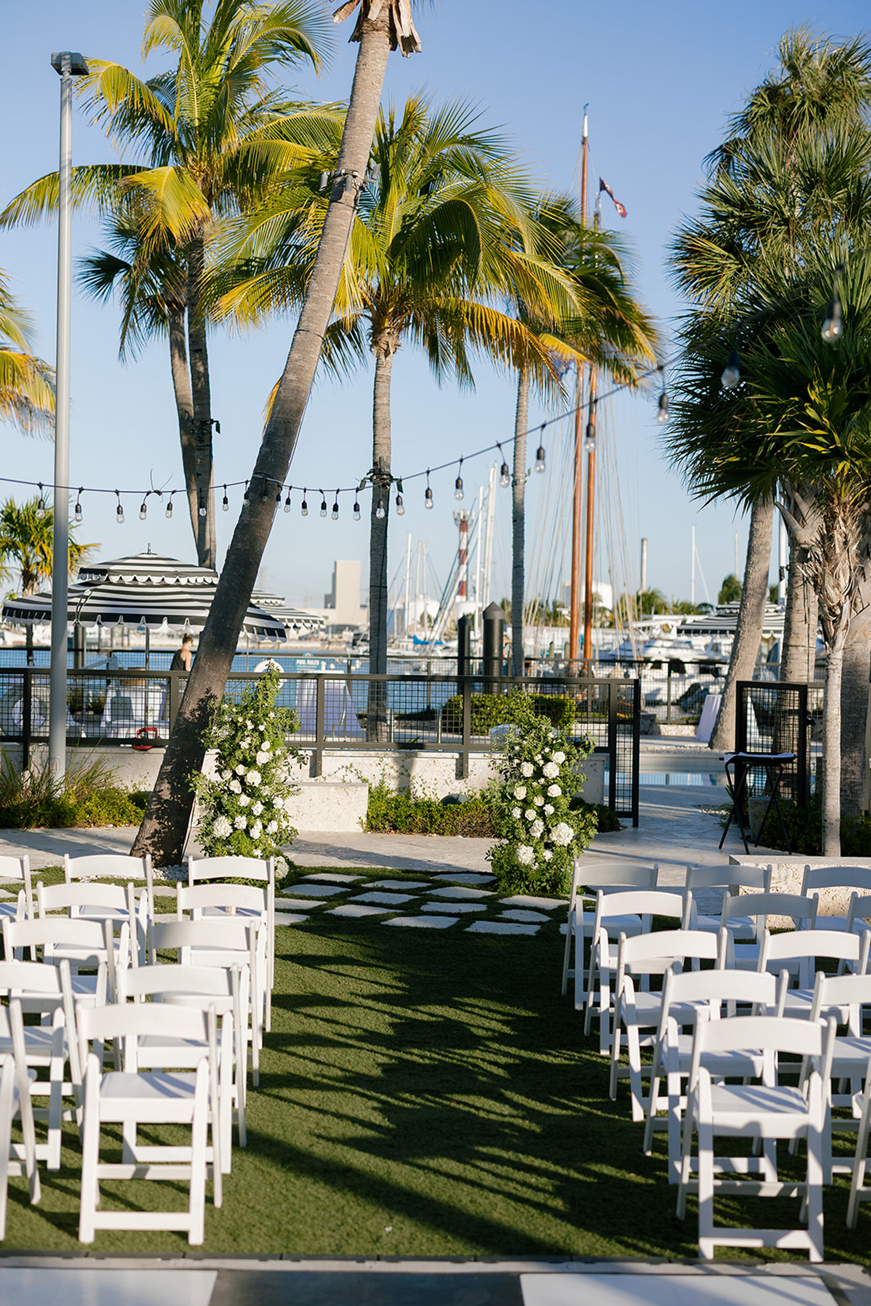 Ceremony setup with white chairs and floral arrangements by palms.