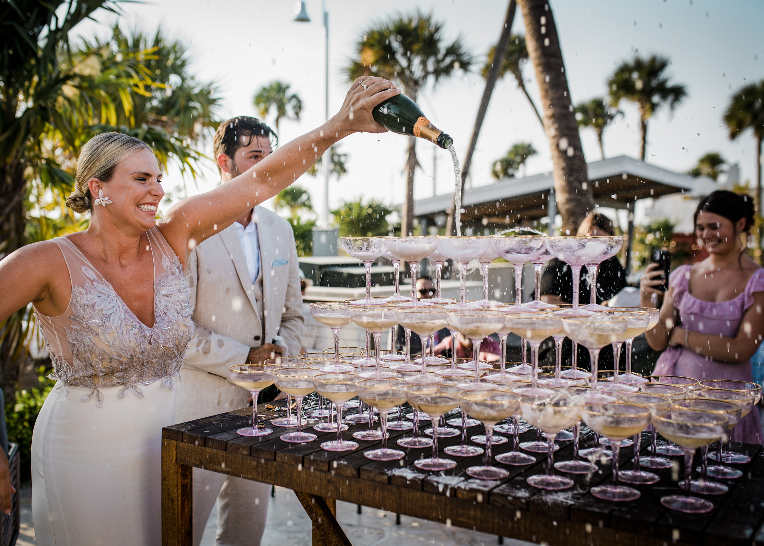 Bride pouring champagne tower outdoors, smiling joyfully.